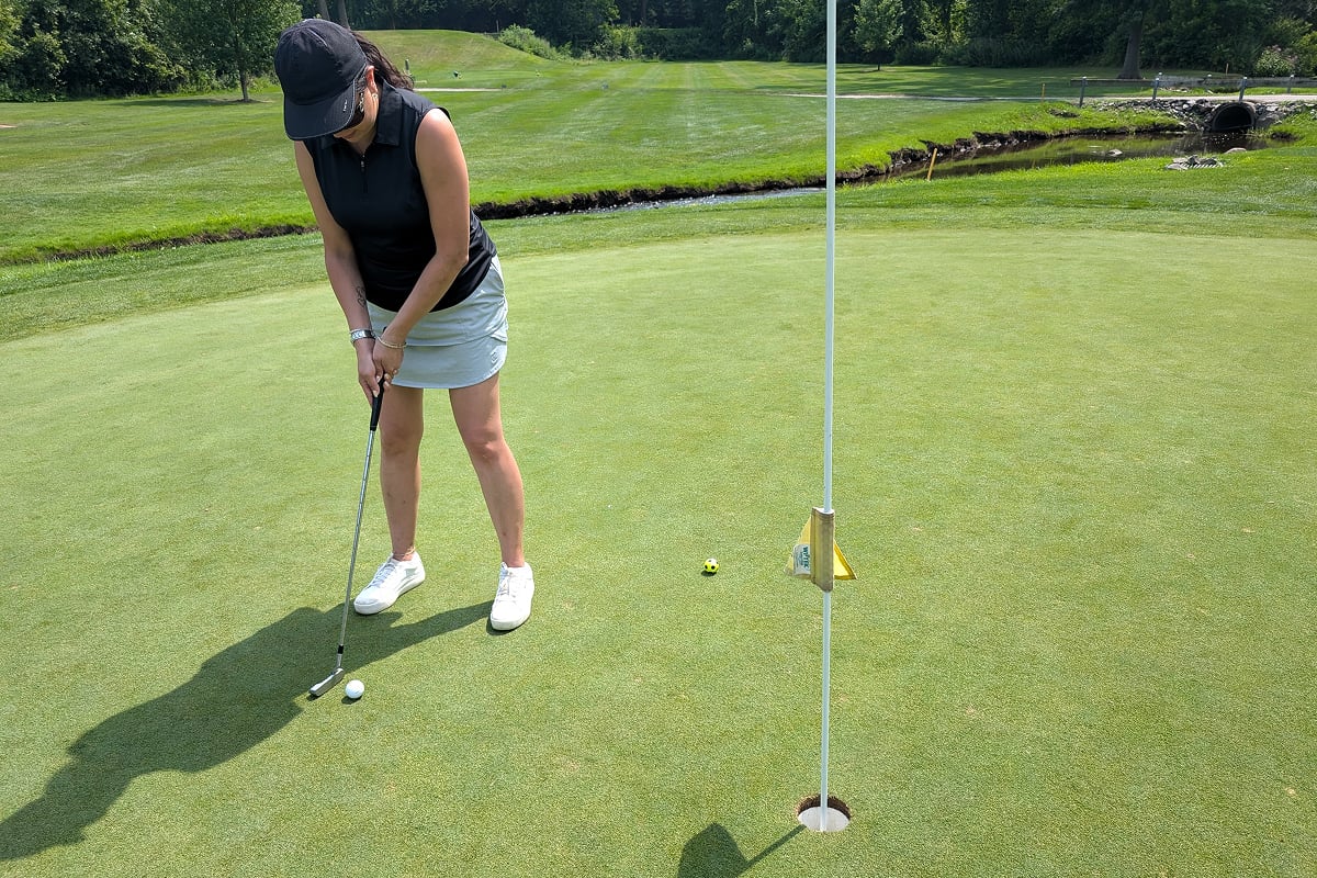 A woman in a black shirt and gray shorts playing golf. She is on the green with a ball and a putter.