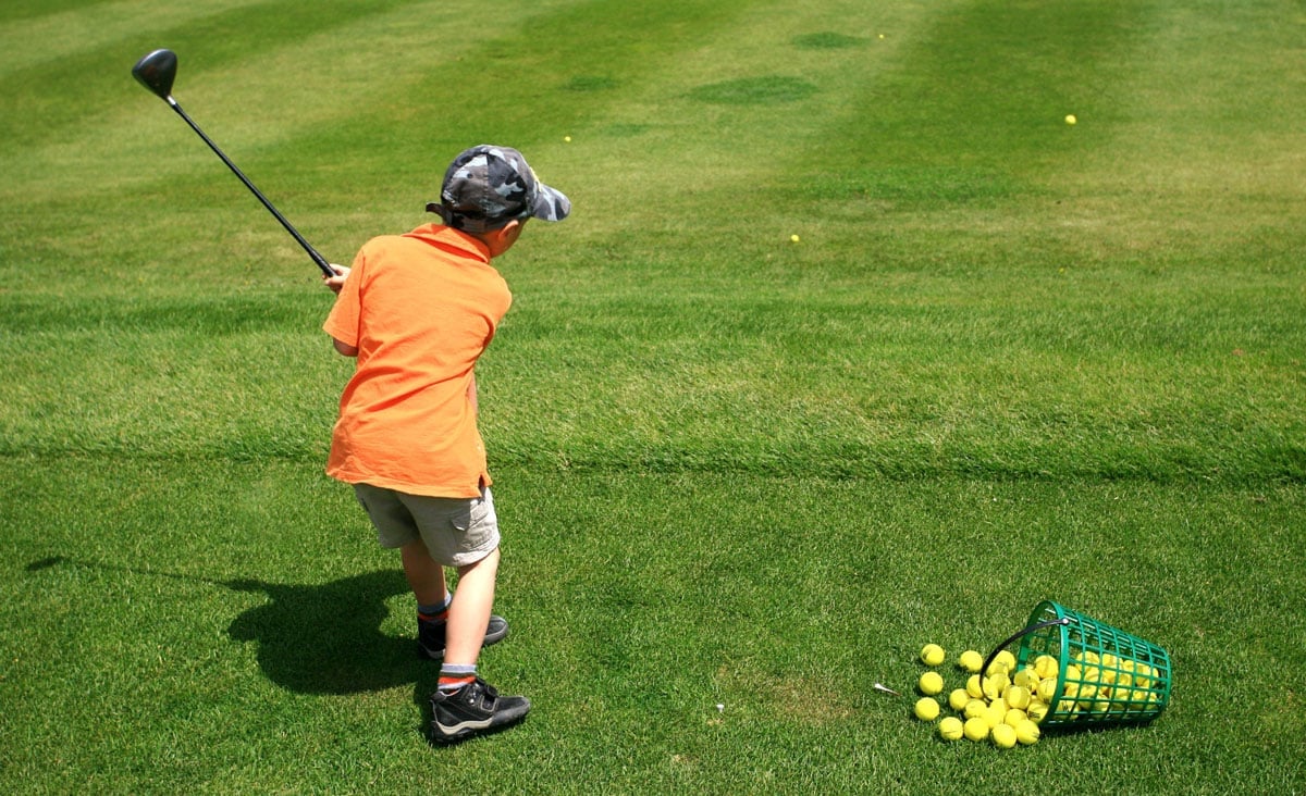 A young boy in an orange shirt swings a golf club. He is standing on a golf course with a basket of balls.
