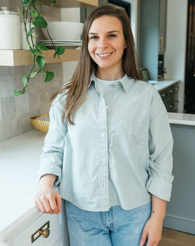 A woman wearing a light blue shirt and jeans in a kitchen. She is standing next to a white counter with a plant hanging down.