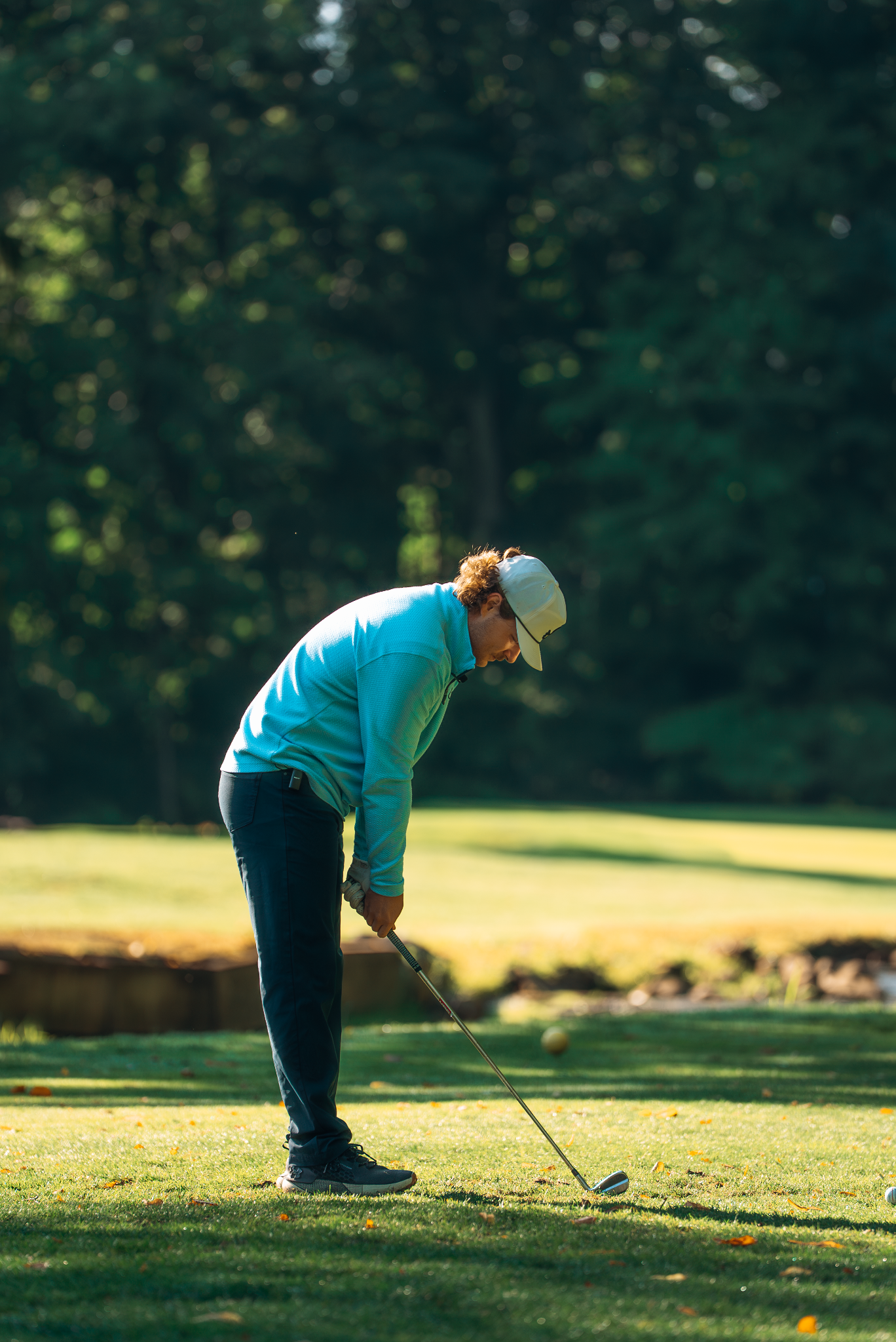 Man in a blue shirt swinging a golf club on a green course. He is wearing a white cap.