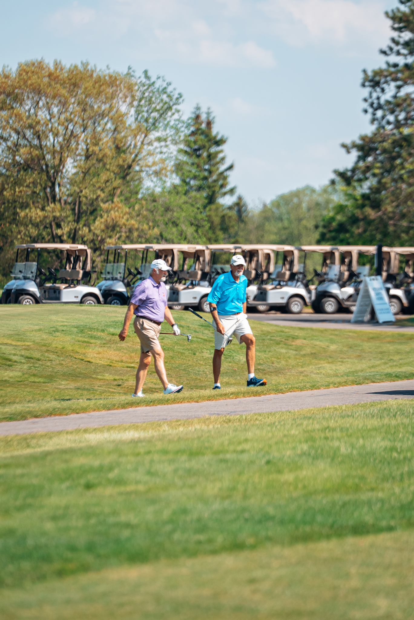 Two golfers walking on a golf course. Several golf carts are parked in the background.