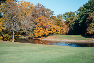 A serene autumn landscape featuring a manicured golf course, a calm pond, and vibrant fall foliage.