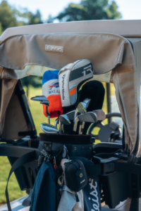 Close-up of a golf cart's beige canopy showing an 'E-CAT' label. Golf clubs and a driver headcover are visible in the background on a sunny day.